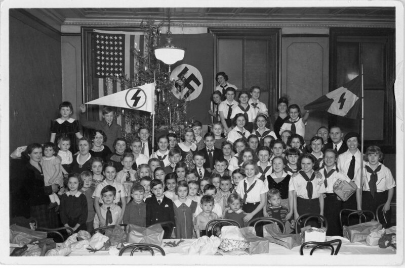 Young boys and girls, not all of them in uniform, crowd together in front of Nazi and American flags. The mood of this image is jubilent with young children smiling in the front.